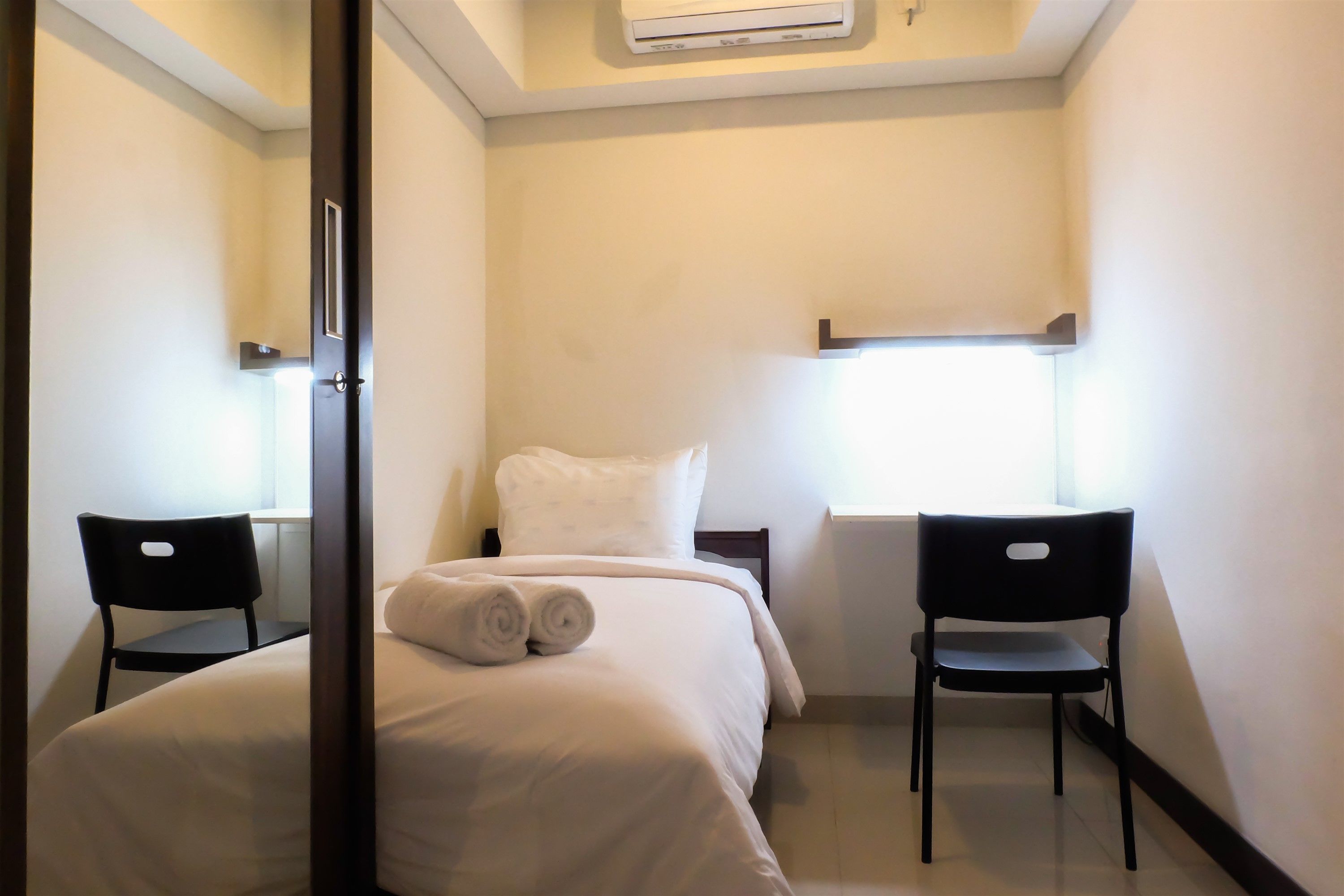 Une chambre d’hôtel avec un lit, deux chaises, une fenêtre et une étagère fixée au mur.