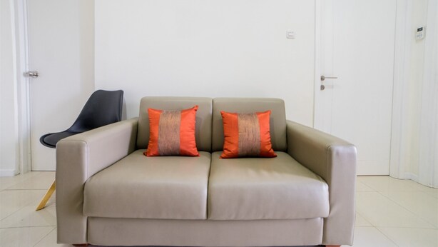 Un sofá beige con dos almohadas naranjas, una silla negra y una puerta blanca en una habitación con paredes blancas y suelo de baldosas.