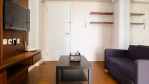 A modern living room with a flat-screen TV mounted on the wall, a black table with a laptop and a bottle, and a purple sofa. - Modern Room Bassura Apartment With Direct Access To Shopping Center (Jakarta)