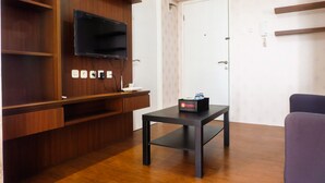 A modern living room with a flat-screen TV mounted on a wooden wall, a black table with a box labeled 'travello', and a door with a handle. - Modern Room Bassura Apartment With Direct Access To Shopping Center (Jakarta)
