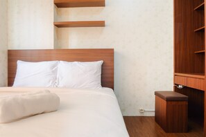 A neatly made bed with white linens and a rolled white towel, a wooden headboard, and a small wooden nightstand with a drawer. - Modern Room Bassura Apartment With Direct Access To Shopping Center (Jakarta)
