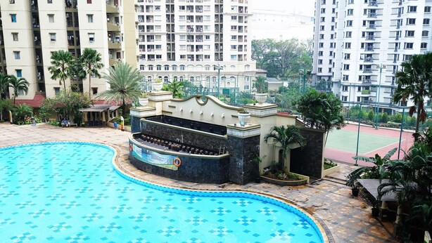 A swimming pool with a tiled pattern, surrounded by a stone deck. There is a small structure with a sign, possibly a poolside bar or lounge area, and a tennis court in the background.