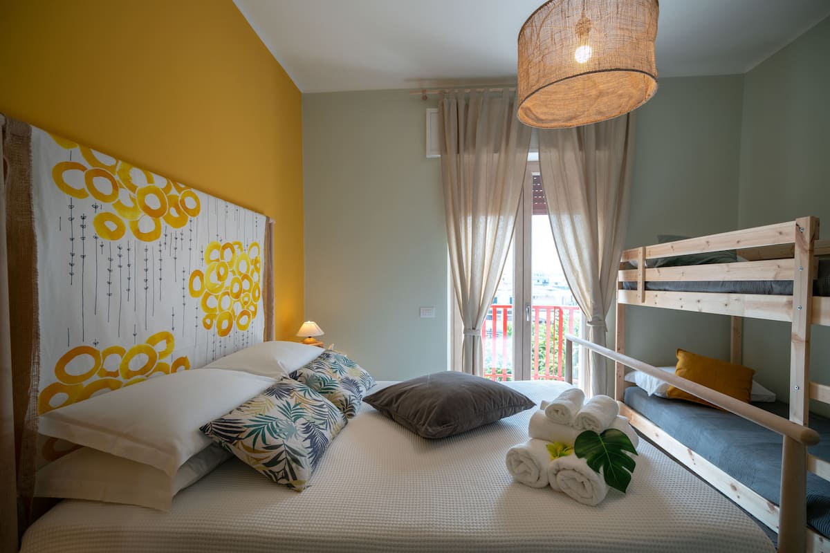 Een slaapkamer met een stapelbed, een groot bed met veel kussens, een hanglamp en een raam met gordijnen.