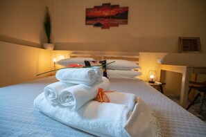 Una cama bien hecha con toallas blancas enrolladas y una cinta naranja pequeña, un cuadro colgado en la pared, y una mesita de noche con una lámpara.