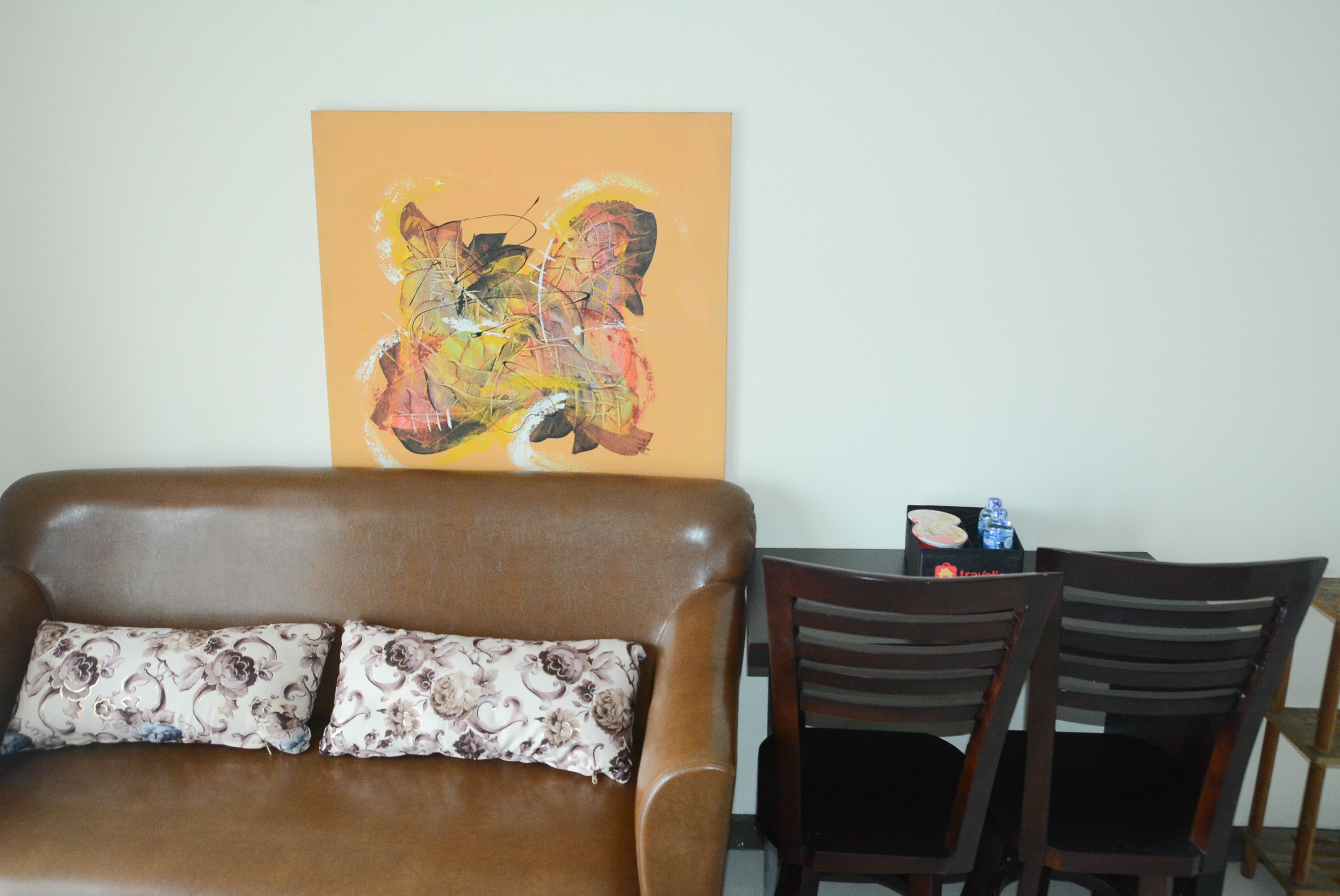 Una sala de estar con un sofá de cuero marrón, dos almohadones estampados, una mesa de comedor de madera con sillas y un cuadro abstracto de colores en la pared.