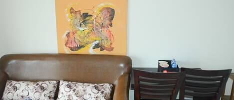 Una sala de estar con un sofá de cuero marrón, dos almohadones estampados, una mesa de comedor de madera con sillas y un cuadro abstracto de colores en la pared.