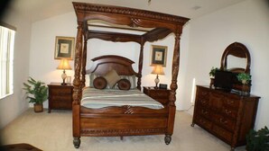 A four-poster bed with a wooden frame and a matching headboard, flanked by two nightstands with lamps and framed pictures. A wooden dresser with a mirror and a plant is to the right. - Ip60402 - Sand Hill - 5 Bed 3 Baths Villa (Davenport)