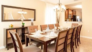 Une salle à manger avec une grande table rectangulaire dressée pour six personnes, entourée de six chaises. Un lustre est suspendu au plafond, et un grand miroir est fixé au mur au-dessus de la table.