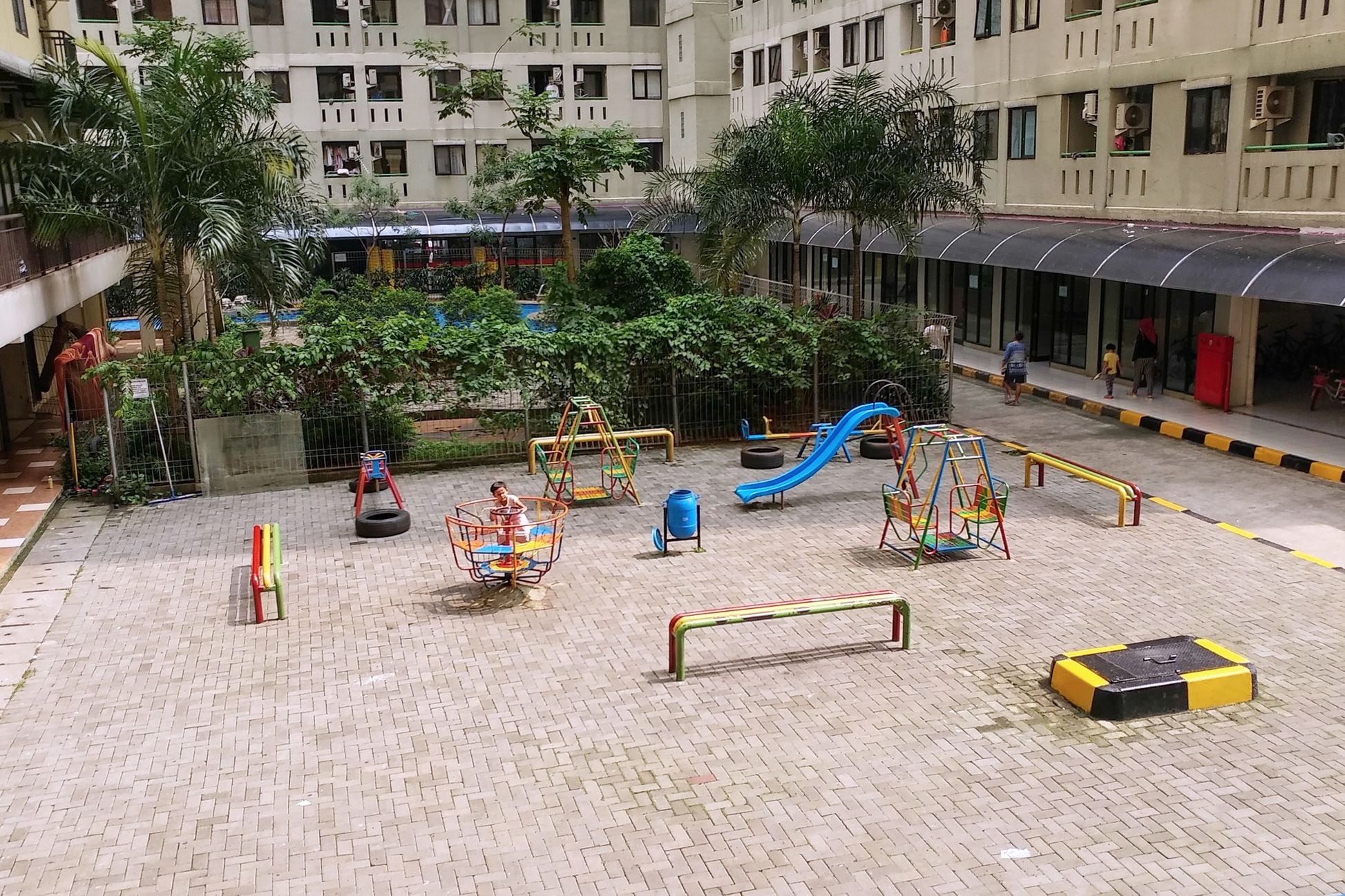 Un parque infantil al aire libre con diversas estructuras de juego, rodeado de pavimento de ladrillo y follaje.