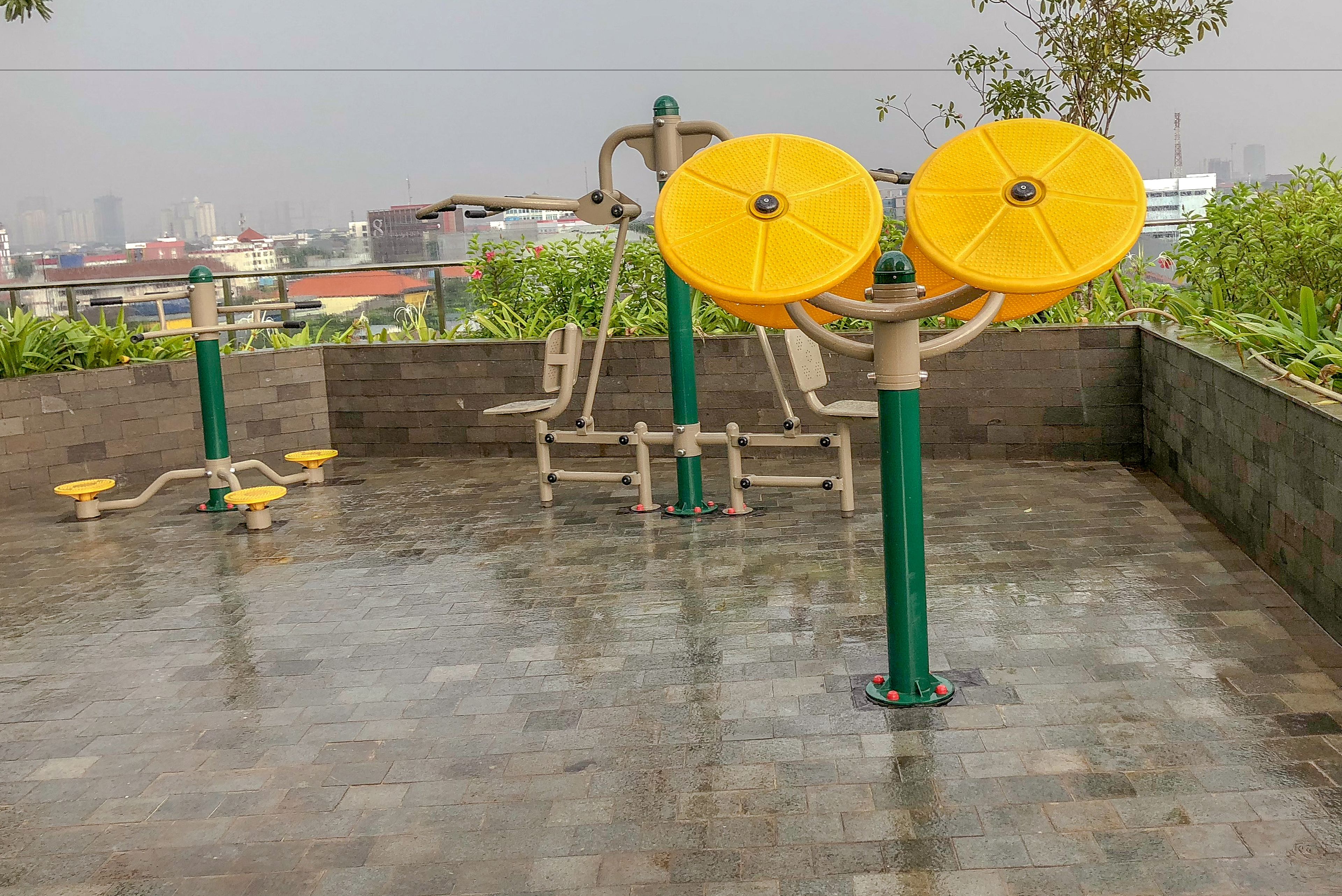 An outdoor exercise area with various fitness equipment, including a yellow circular machine and a multi-directional machine, set against a backdrop of buildings and greenery.