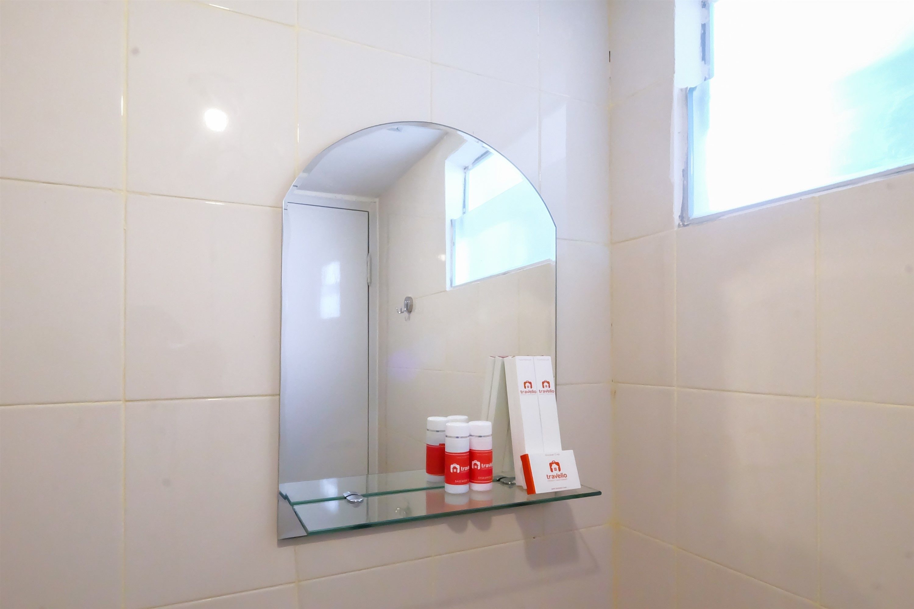 A bathroom with white tiled walls, a glass shelf with toiletries, and a mirror reflecting the room.