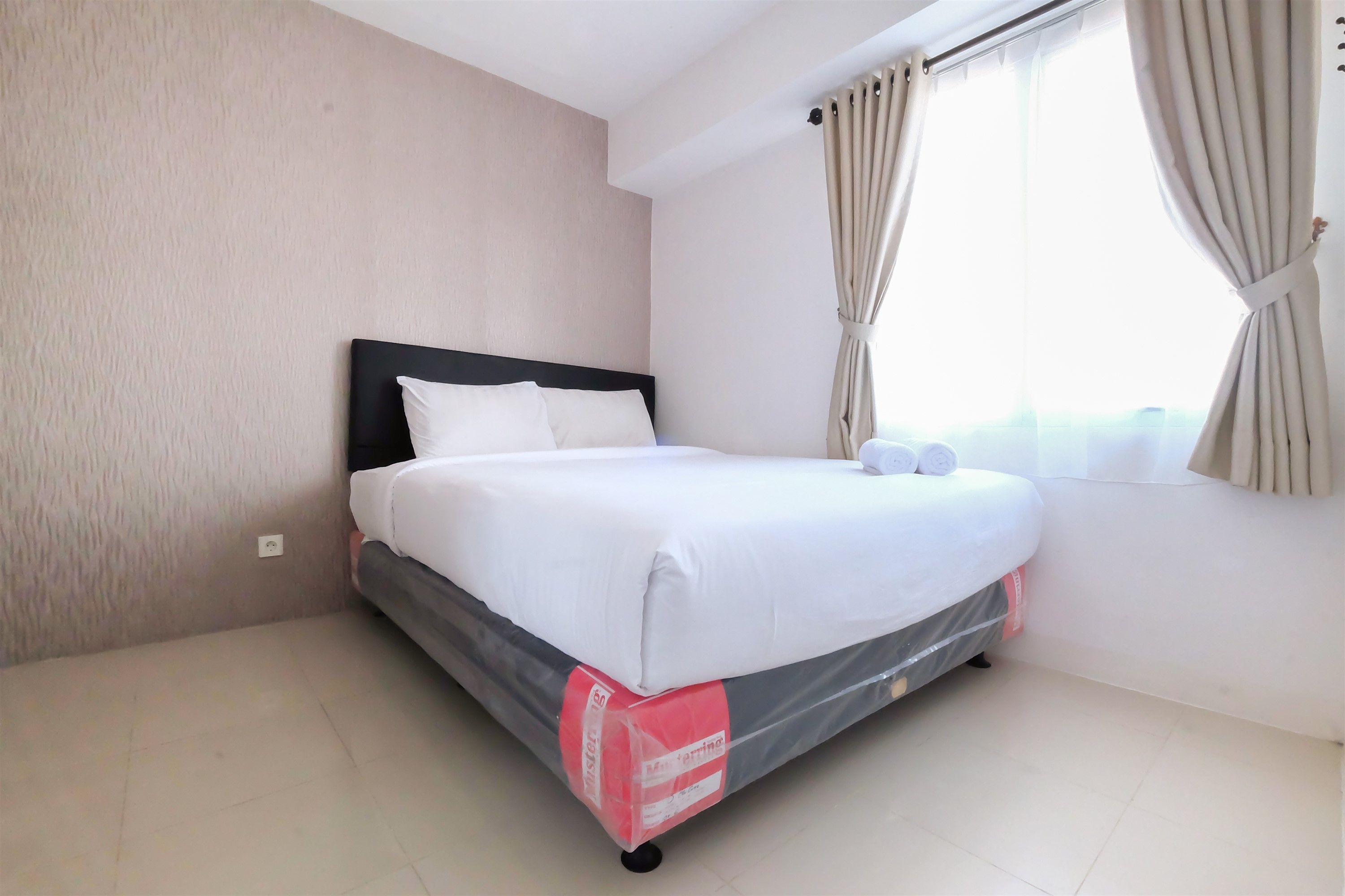A neatly made bed with white linens, a black headboard, and a window with beige curtains.