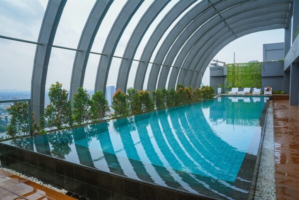 A rooftop infinity pool with a curved glass ceiling, surrounded by greenery and a city view.