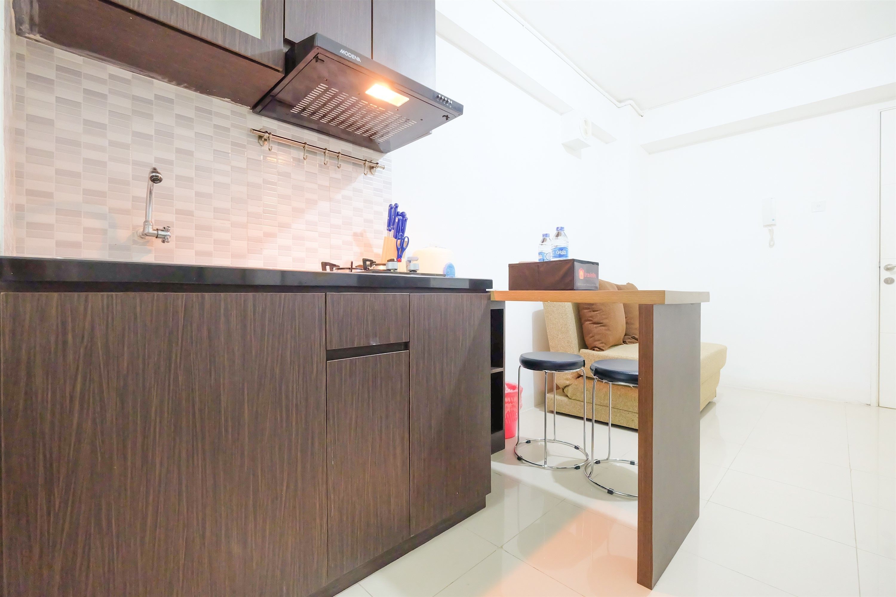 A modern kitchen with dark wood cabinets, a tiled backsplash, and a built-in stove. There is a dining area with a table and chairs, and a sofa in the background.