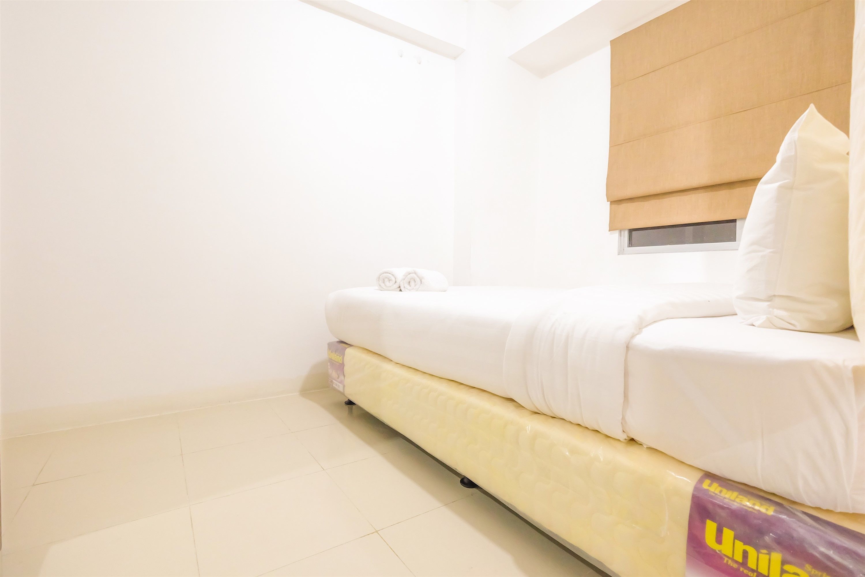 A small, bright room with a bed, a window with a blind, and a tiled floor.