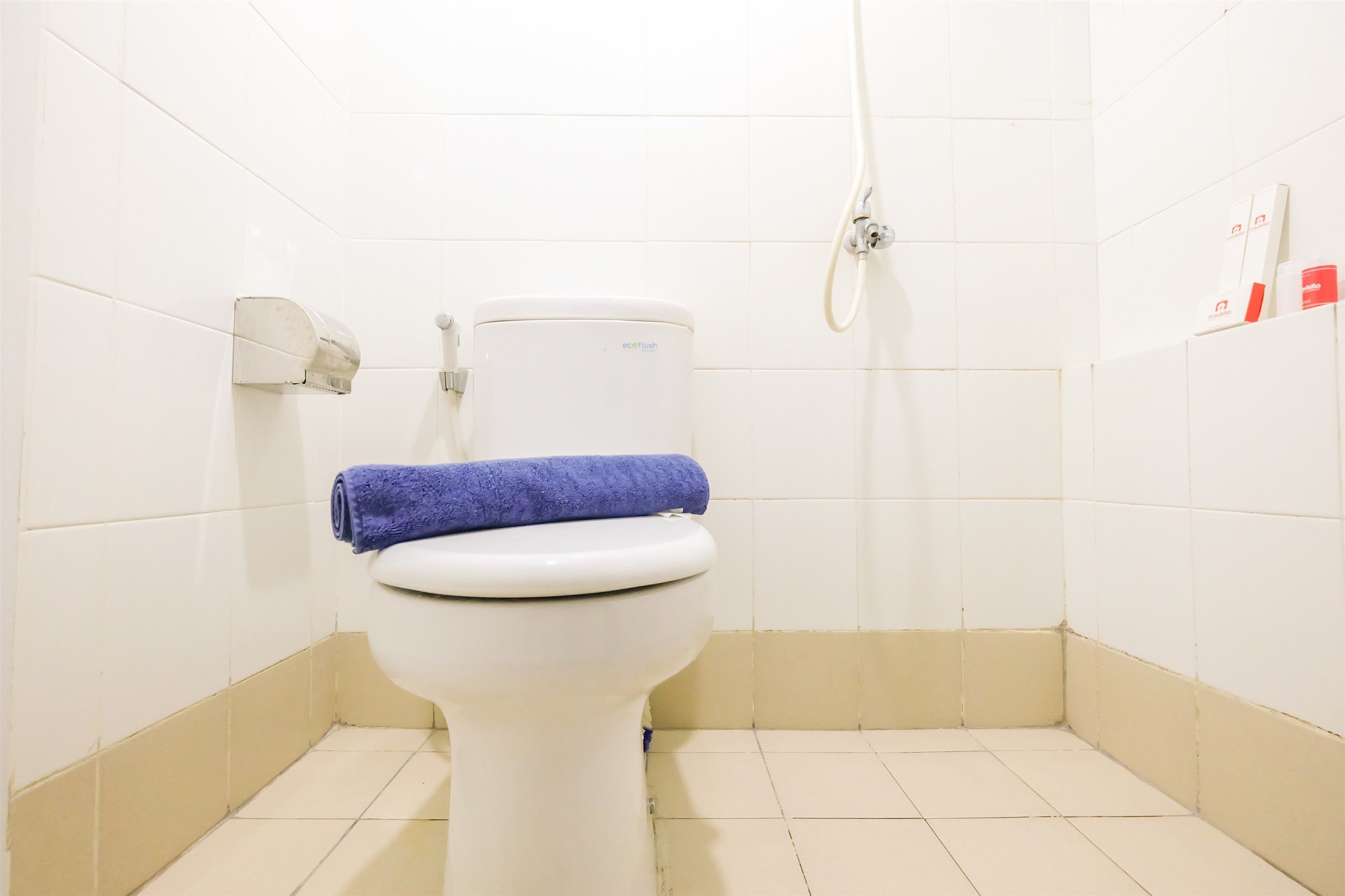 A white toilet with a blue towel on top, a hand dryer, and a shower hose in a bathroom with white tiles.