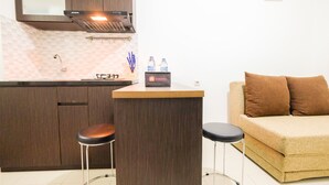 A compact kitchen with a built-in stove, a microwave, and a coffee machine. There are two stools and a small table with a water bottle and a travel bag on it. A beige sofa is positioned next to the kitchen. - Enjoy Jakarta! Bassura City Apartment (Jakarta)