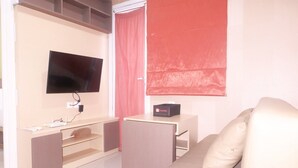 A room with a flat-screen TV mounted on the wall, a small desk with a computer and a box labeled "Travelio," a sofa, and a window with a red blind.