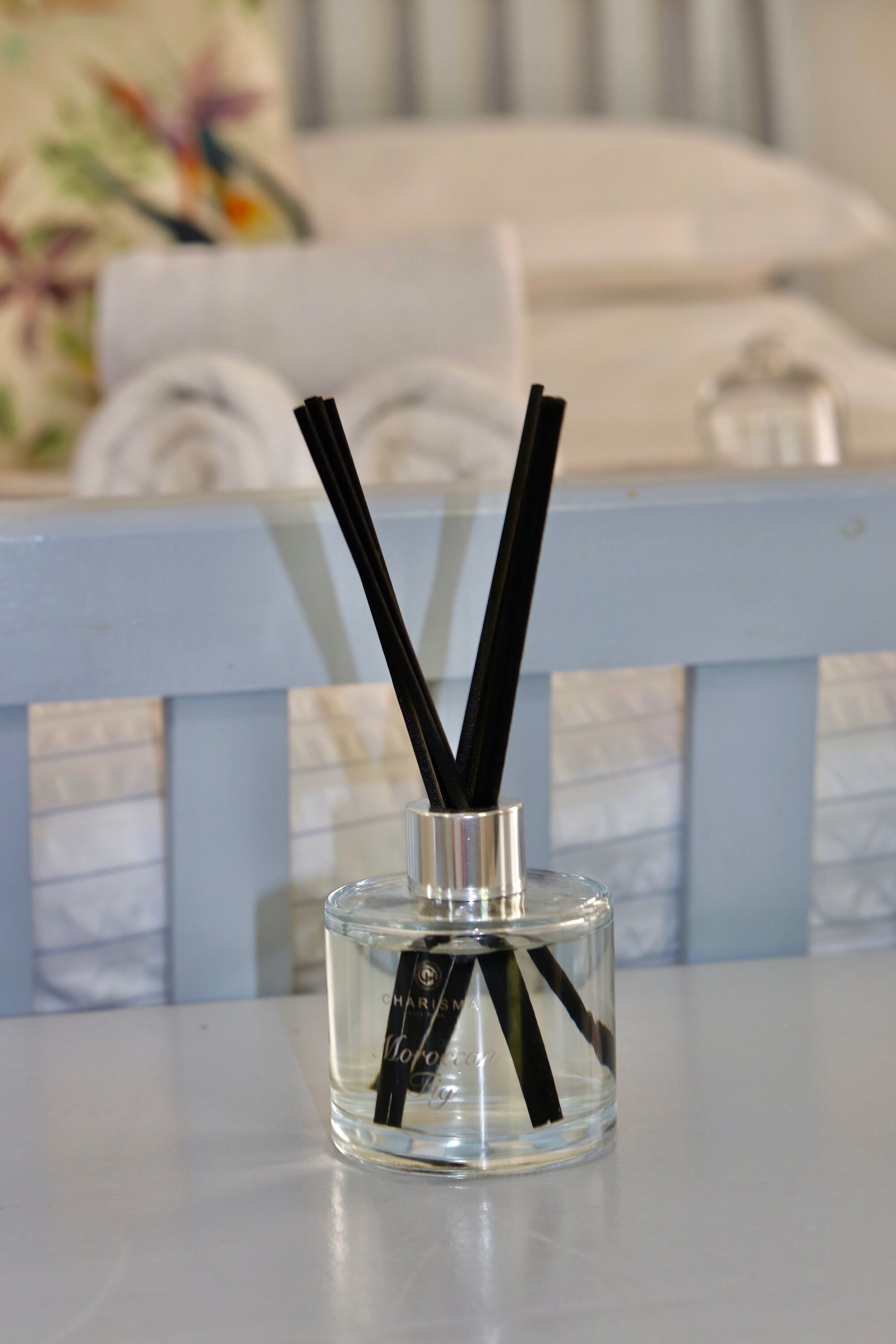 A clear glass bottle with black reeds and a label, placed on a table with a blurred background of a bed and pillows.
