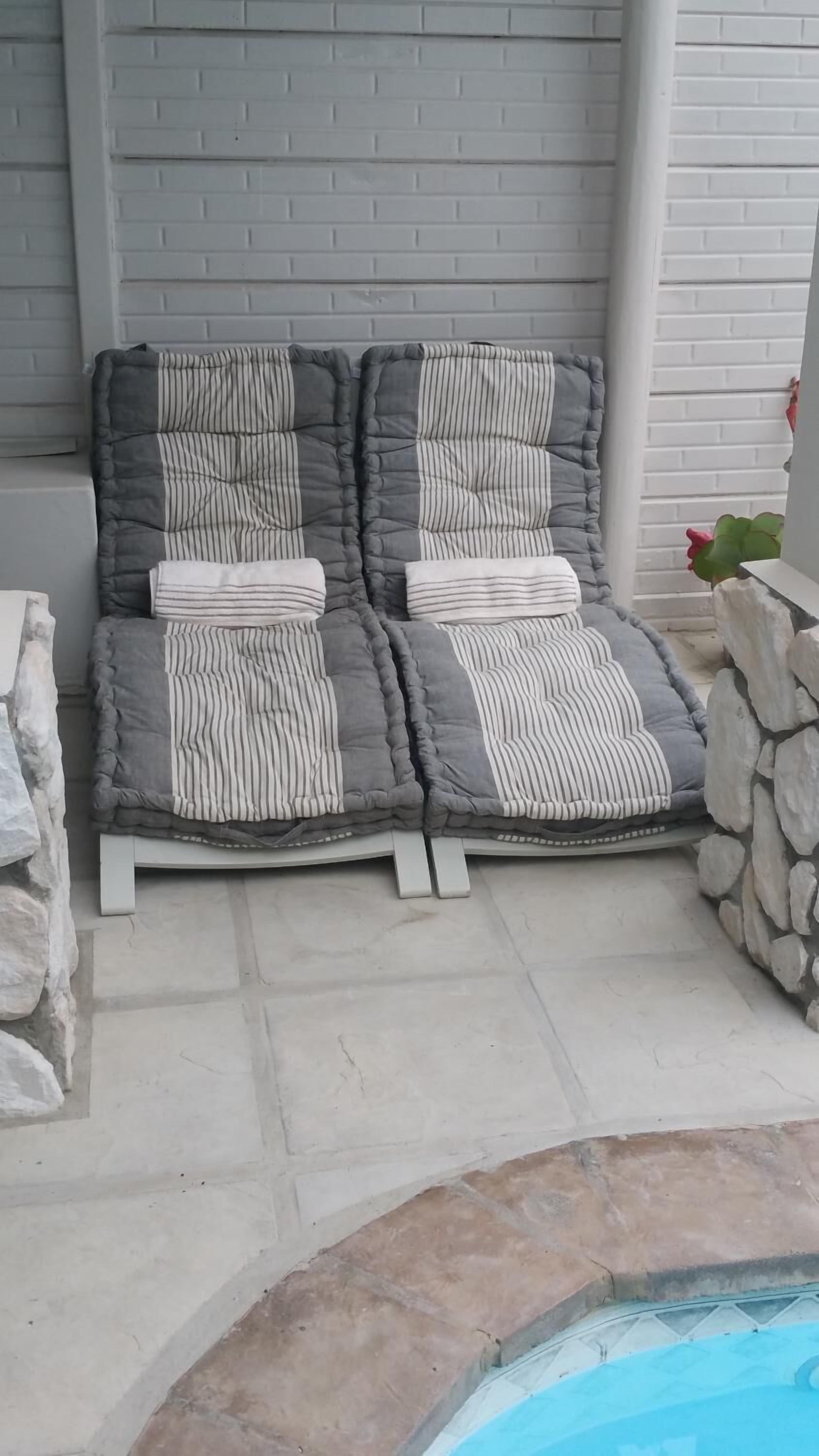 A two-seater lounge chair with striped cushions, positioned on a stone-tiled patio adjacent to a pool.
