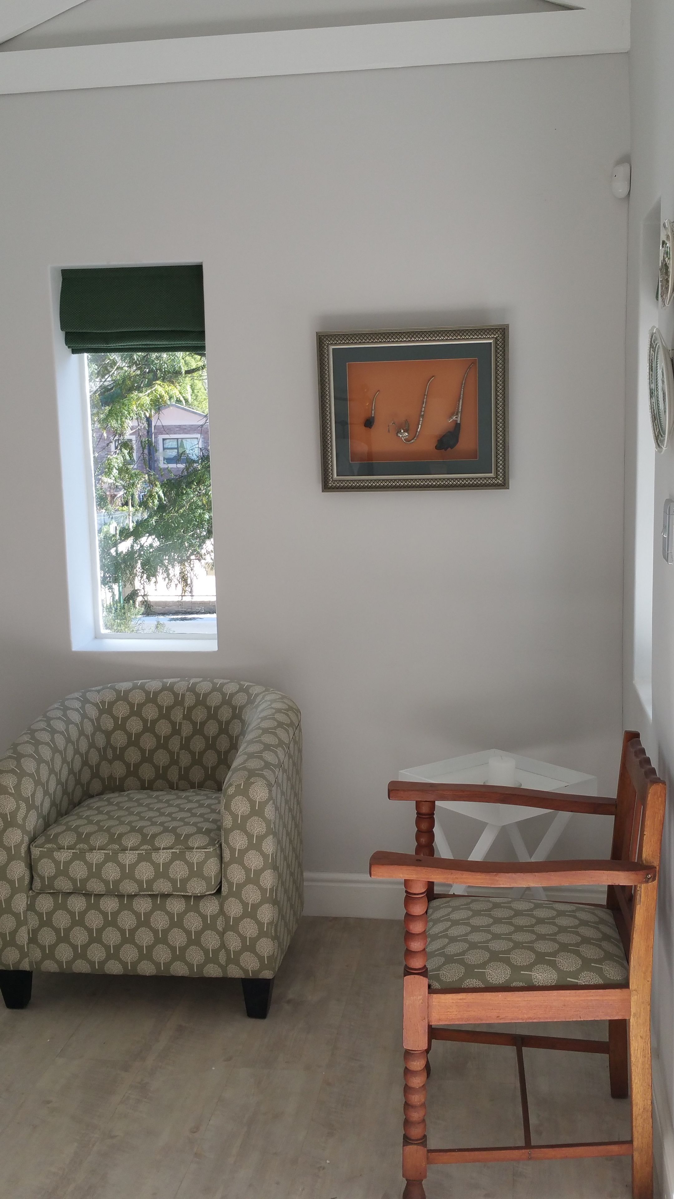 Une pièce avec un canapé à motifs, une chaise en bois, un tableau encadré et une fenêtre donnant sur des arbres et des immeubles.