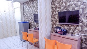 A hotel room with a large flat-screen TV mounted on the wall, a wooden desk with a computer and a coffee machine, and two orange chairs. - Warm Cozy Menteng Square Apartment (Jakarta)