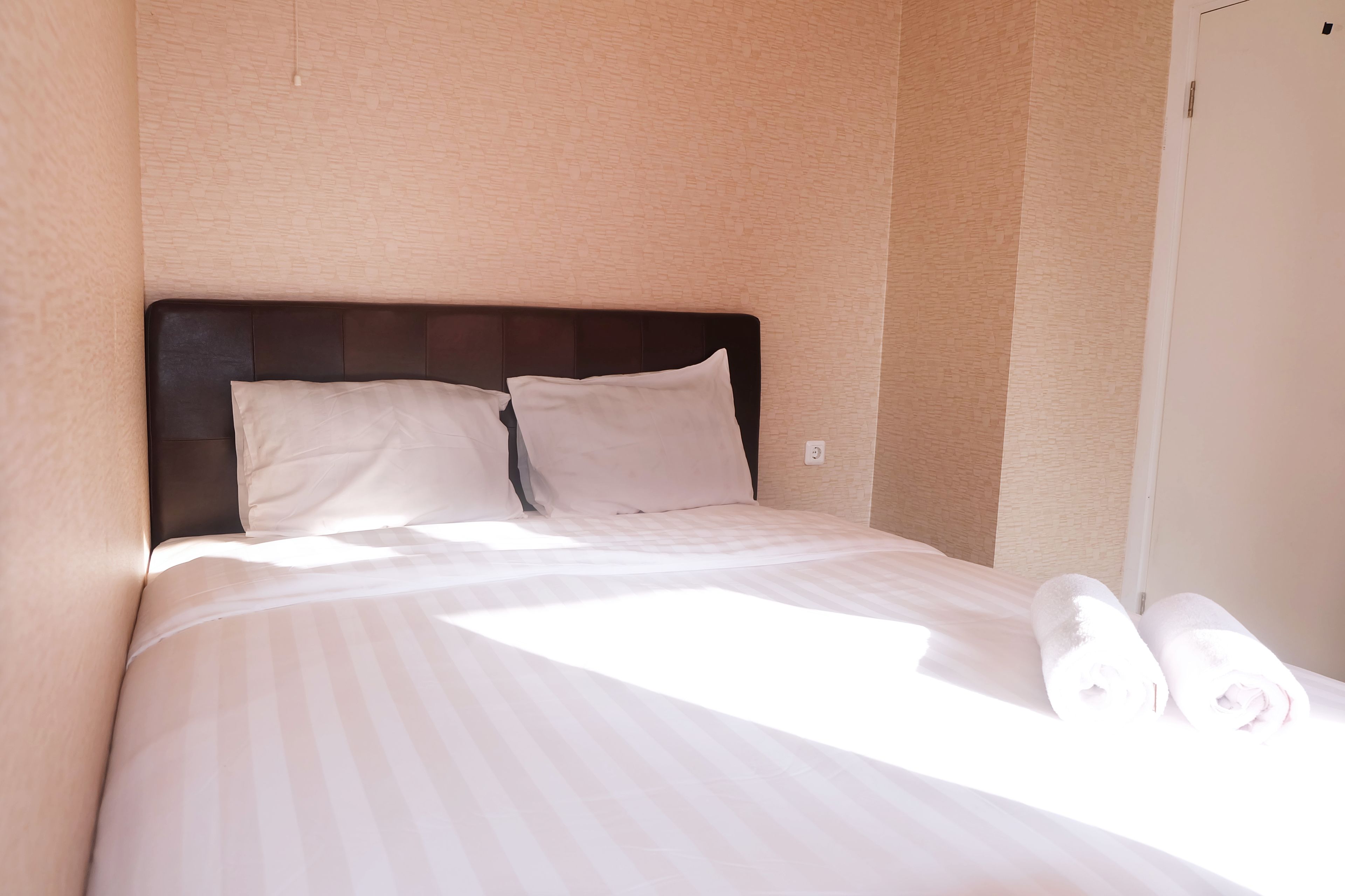 A neatly made bed with white linens and two pillows, a dark headboard, and a pair of rolled towels on the side.