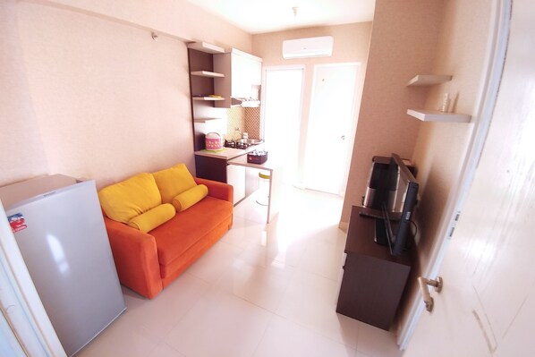 A compact living space with a kitchenette, a sofa with yellow cushions, a refrigerator, and a door leading to an adjacent room.