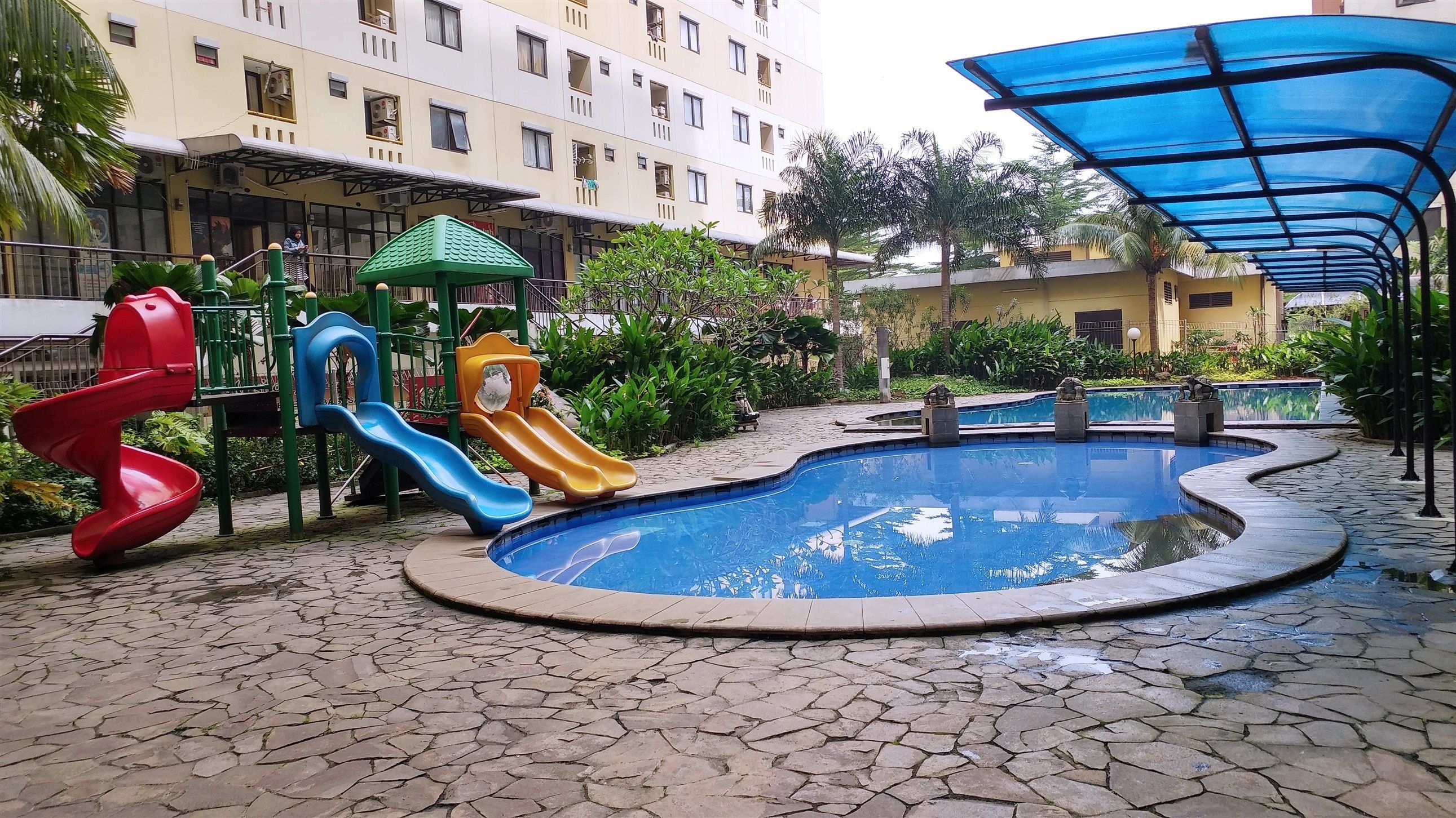 Ein Spielplatz mit Rutschen und einem kleinen Pool, umgeben von einer gepflasterten Fläche und Wohngebäuden.