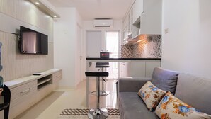 A modern living room with a grey sofa, a flat-screen TV mounted on the wall, a kitchen area with a tiled backsplash, and a bar stool. - Minimalist Bassura Apartment Direct Access to Bassura City Mall (Jakarta)