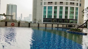 Una piscina con agua cristalina, rodeada por un muro bajo y una barandilla. Al fondo, edificios modernos con ventanas amplias y balcones.