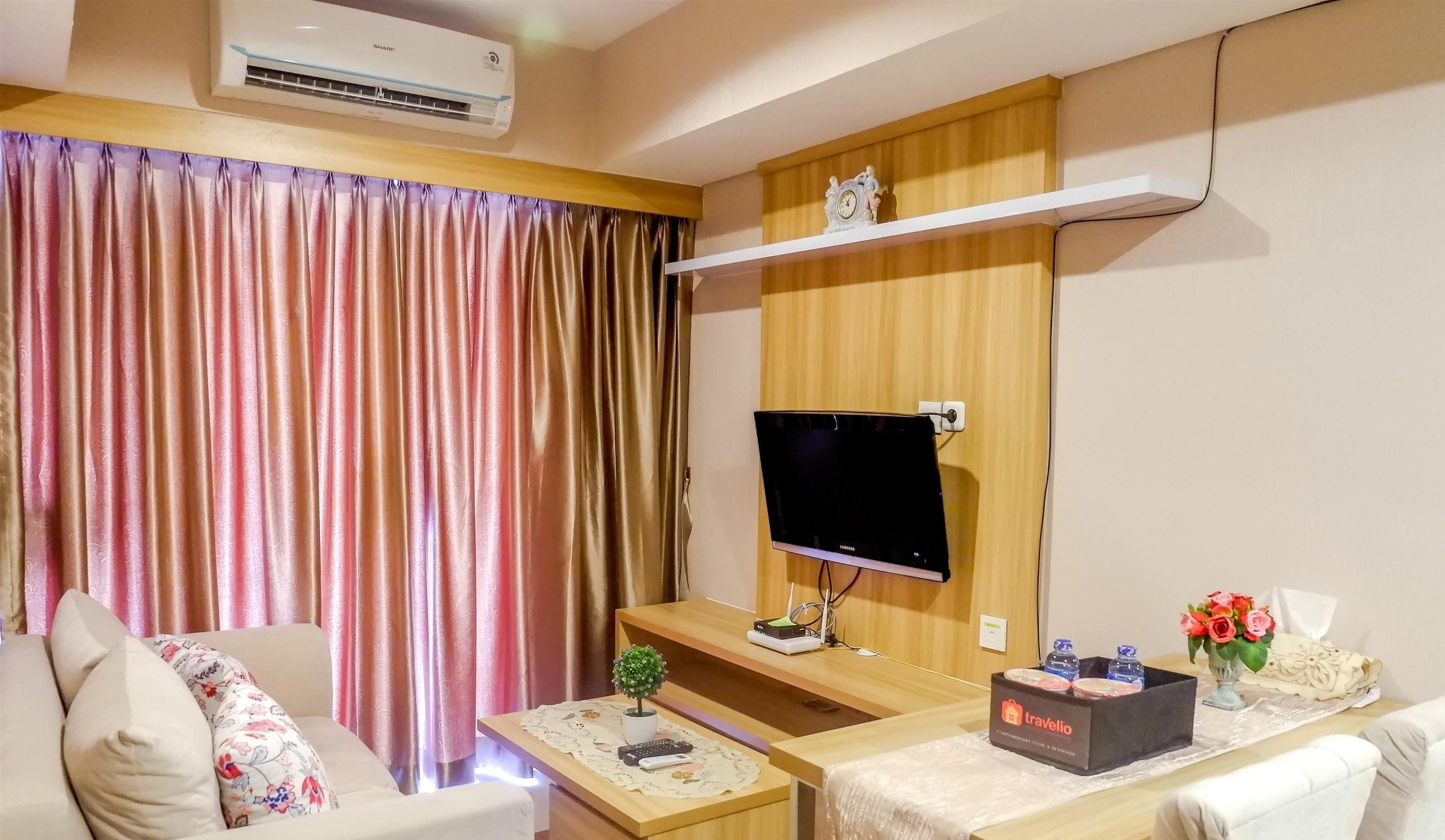A hotel room with a flat-screen TV mounted on a wooden wall, a sofa with floral cushions, a small table with a remote control, and a tray table.