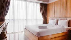 Hotelkamer met houten hoofdeinde, een groot bed met wit beddengoed, een bureau met stoel en dunne gordijnen voor het raam.
