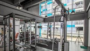 Een sportschool met gewichthefapparatuur, banken en uitzicht op een gebouw met blauwe ramen.