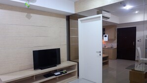 Un salon moderne avec un téléviseur à écran plat posé sur un meuble en bois, une porte en verre donnant sur la cuisine et une porte blanche.