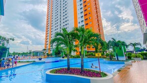 A swimming pool surrounded by palm trees and a high-rise building in the background. - Comfy Studio Apartment @ Green Pramuka City (Jakarta)