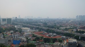 A cityscape with a mix of high-rise buildings and smaller structures, a busy road with vehicles, and a clear sky. - Comfy Studio Apartment @ Green Pramuka City (Jakarta)