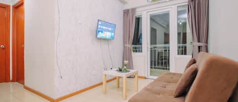 A living room with a sofa, a television mounted on the wall, a small table with a plant, and a sliding glass door leading to a balcony.