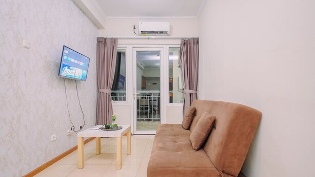 A living room with a brown sofa, a small table with a plant, and a mounted TV displaying a movie. - Apartment Palazzo/Grand Palace Kemayoran (Jakarta)