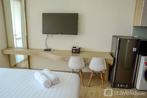 A hotel room with a bed, a TV mounted on the wall, a desk with two chairs, a refrigerator, and a mirror.