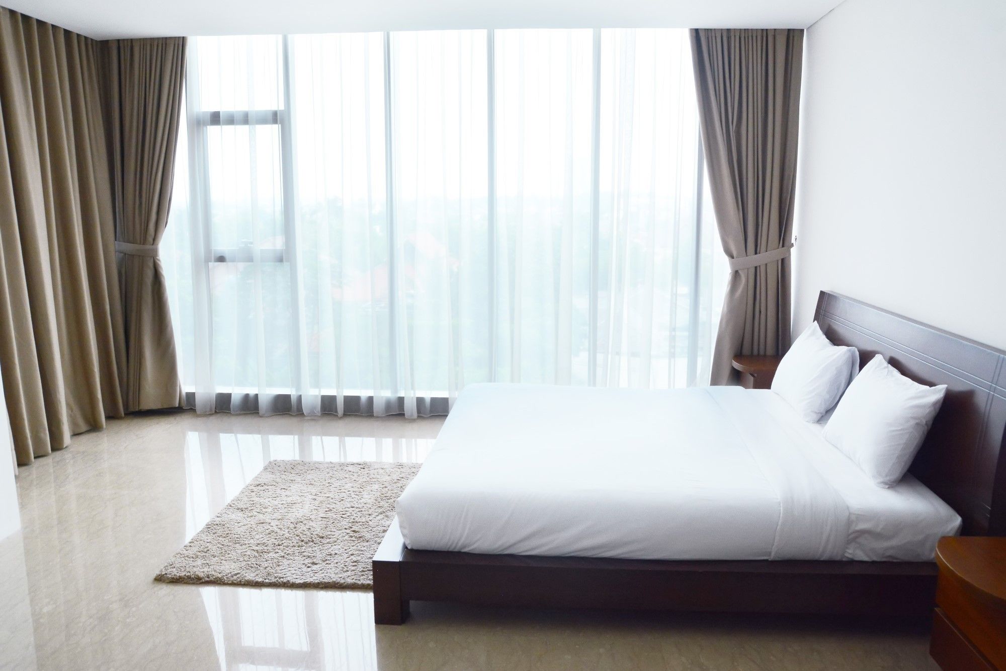 A modern hotel room with a large bed, a wooden headboard, and a view of the city through sheer curtains.