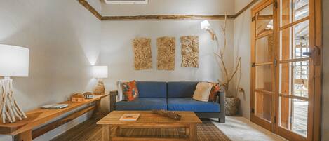 A living room with a blue sofa, wooden coffee table, and a bench. There are three wall decorations, a lamp, and a window with wooden shutters.