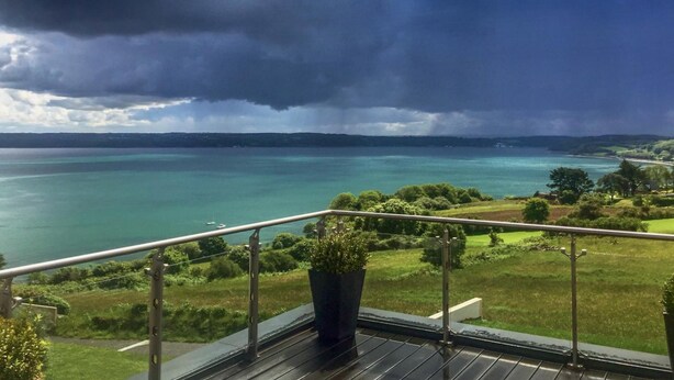 Un balcone con vista su un lago e dolci colline lontane, sotto un cielo nuvoloso.