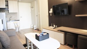 Ein modernes Wohnzimmer mit einer Couch, einem Couchtisch mit Gegenständen, einer Küchenzeile inklusive Kühlschrank und Spüle sowie einem an der Wand montierten Fernseher.