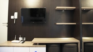 Ein modernes Hotelzimmer mit einem an der Wand befestigten Flachbildfernseher, einem Holztisch mit Telefon und Router sowie einer kleinen Sitzecke mit gepolstertem Sitz.