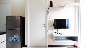 Une pièce de taille modeste avec une porte blanche, un réfrigérateur en acier inoxydable, un téléviseur à écran plat fixé sur une étagère blanche et un tapis rouge sur le sol.