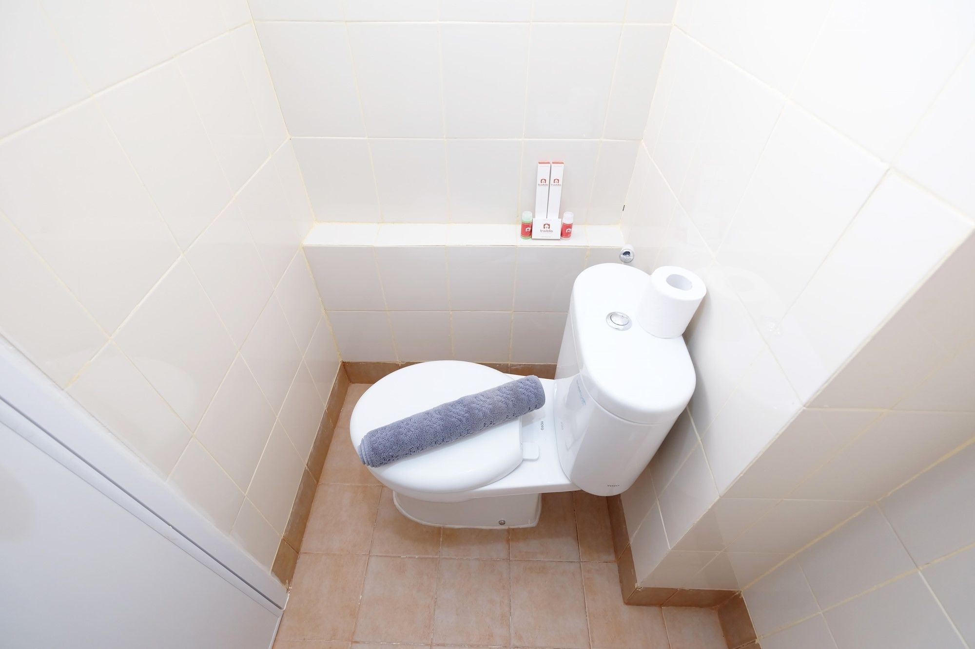 A small, compact bathroom with a white toilet, a towel on the seat, and a small shelf with toiletries.