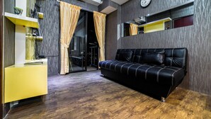 A modern living room with a black leather sofa, wooden flooring, a yellow side table, and a glass door leading to a balcony. - Best Price Tamansari Panoramic Apartment with Mountain View (Bandung)