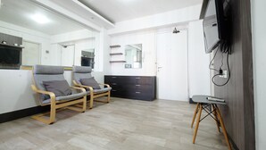 Quarto moderno com duas cadeiras acolchoadas cinzentas, um armário de madeira escura, uma mesinha e uma televisão fixada na parede.