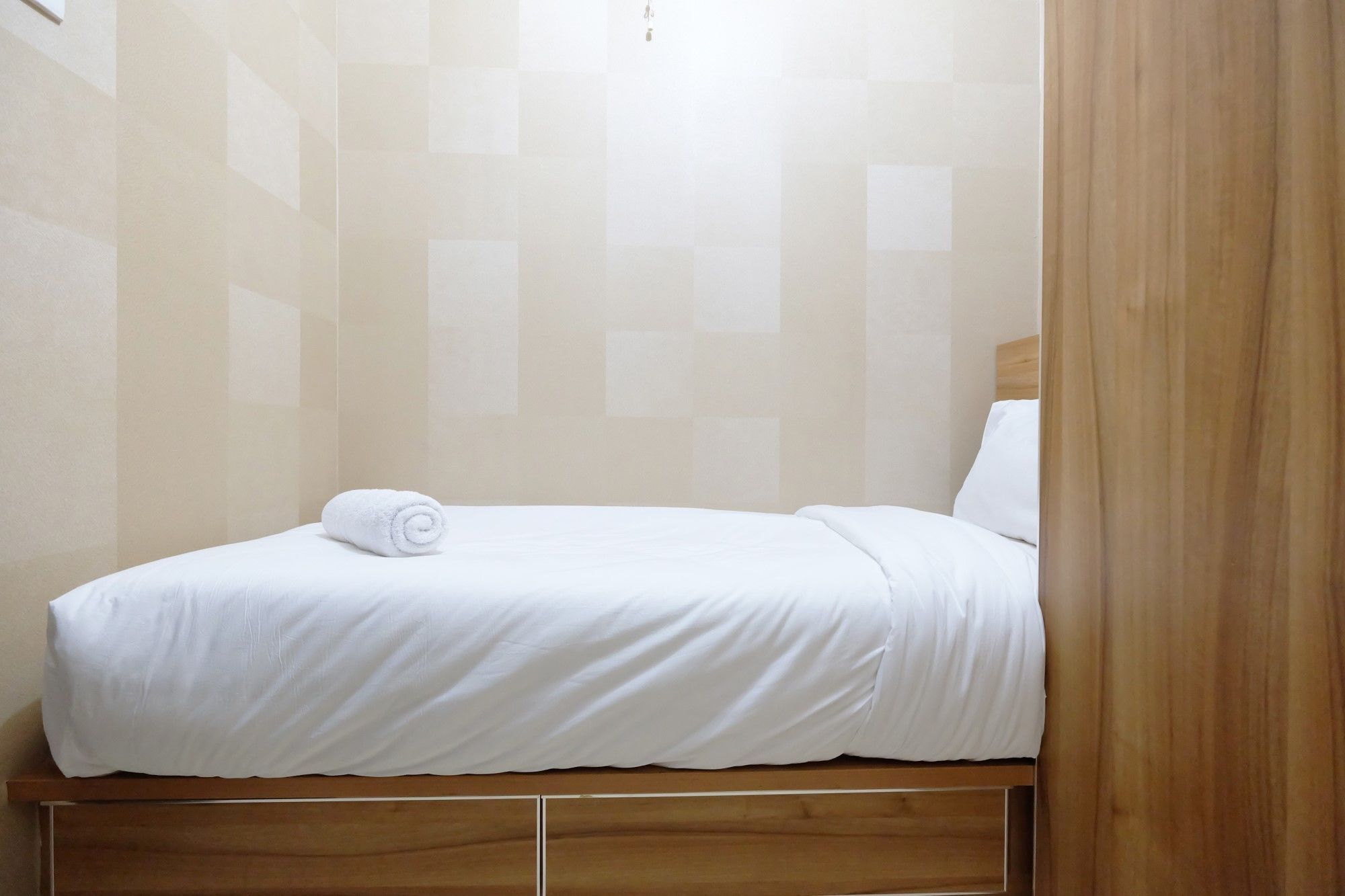 A neatly made bed with white linens and a rolled towel, wooden headboard, and a light-colored tiled wall.